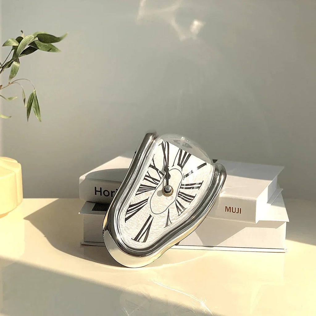 Silver melting clock resting across a stack of two white hardback books on a sunlit desk, with a plant and a candle at the edge of the frame